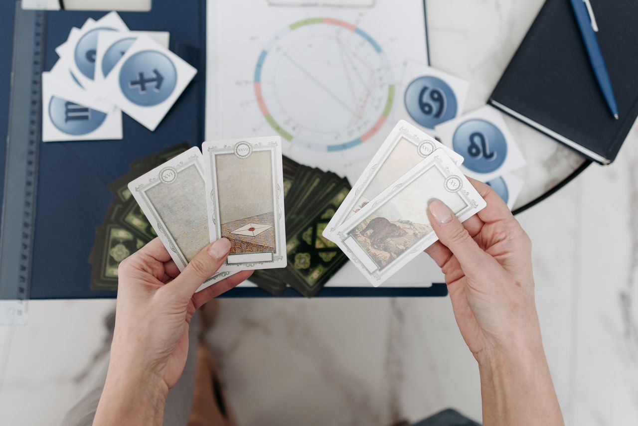 Hands holding tarot cards above an astrological chart with zodiac symbols scattered on the table.