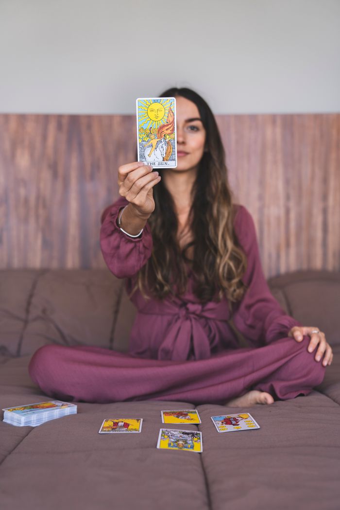 about-01 A woman sitting indoors in a purple dress holding up a tarot card, suggesting a spiritual reading.